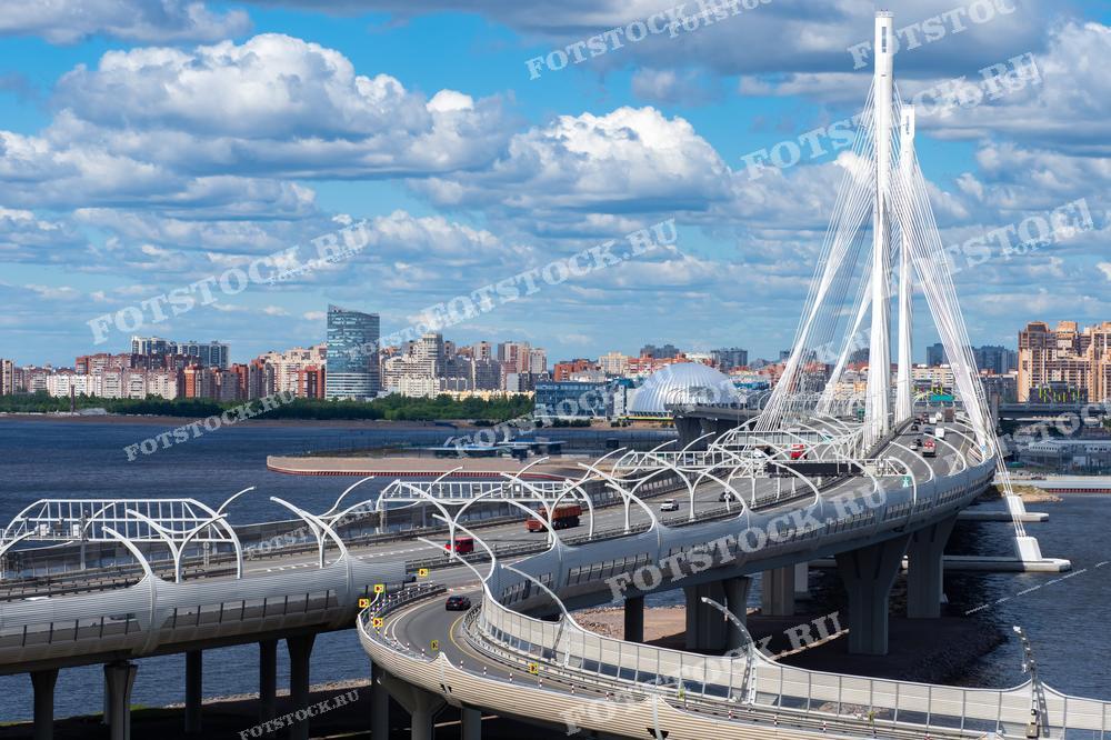 На фотографии изображен Западный скоростной диаметр (ЗСД) и вантовый мост в Санкт-Петербурге. Вид открывается на Финский залив и современные здания на фоне. Летний день с голубым небом и облаками создает живописную картину. ЗСД является важной транспортной магистралью, соединяющей различные районы города.